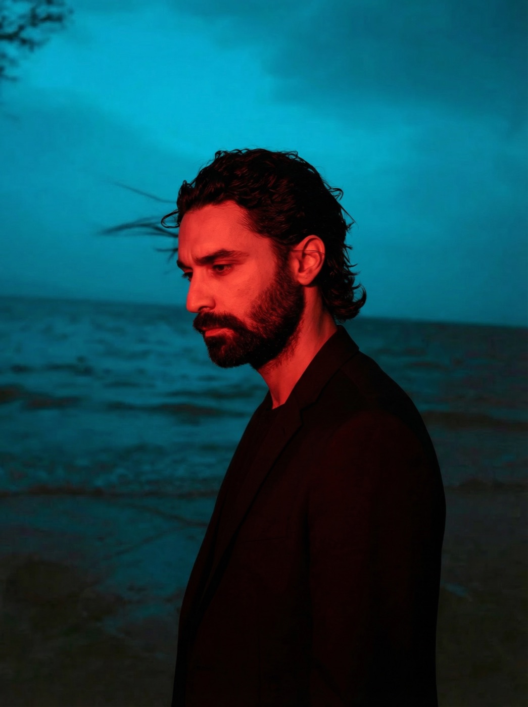 Amos Le Blanc in a dark suit lit red against a teal twilight sea